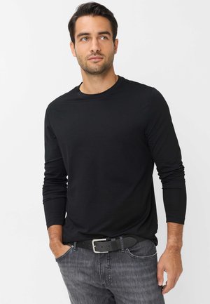 Mand iført sort langærmet skjorte og grå jeans med sort bælte, står med den ene hånd i lommen og kigger mod højre på hvid baggrund.
