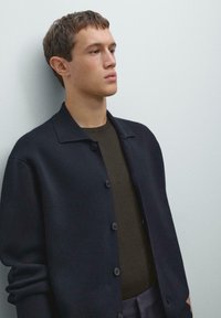 Navy knit cardigan with a collar, button closure, and ribbed texture, layered over a brown shirt, set against a light background.