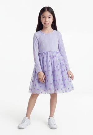 Fille debout portant une robe lavande à manches longues avec une jupe en tulle à pois et des baskets blanches sur fond blanc.