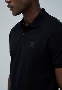 Polo negro con tejido texturizado, un cuello y un sutil parche de logo en el pecho, que presenta un detalle de botón en contraste.
