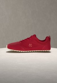 Baskets basses rouges avec lacets à l'avant, semelle texturée et logo subtil de la marque sur le côté, sur fond neutre.