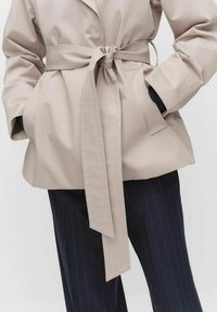 Beżowy płaszcz z paskiem, z zapięciem na guziki i dużymi bocznymi kieszeniami, posiada długie rękawy i luźny krój. Połączony z spodniami w paski.