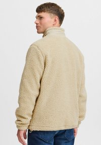 Beige fleece jacket with high collar, textured fabric, and elastic cuffs. Features a drawstring hem and a relaxed fit.