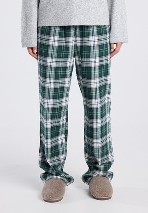 Persoon gekleed in groen en wit geruite broek, grijze trui en beige fuzzy slippers, staand op een witte vloer.