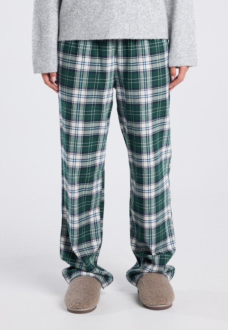 Persoon gekleed in groen en wit geruite broek, grijze trui en beige fuzzy slippers, staand op een witte vloer.