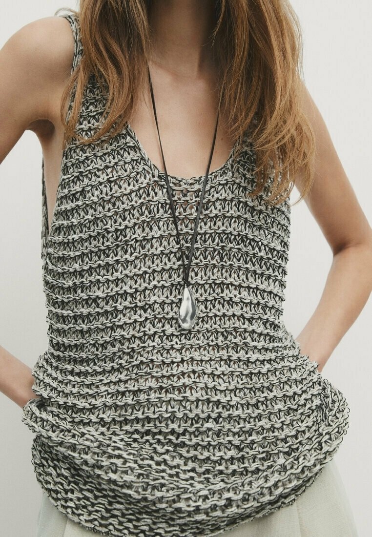 Femme portant un haut gris en tricot épais sans manches et un long collier noir avec un grand pendentif en argent, les mains posées sur les hanches.