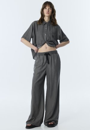 Vrouw staand met handen in de zij, draagt een losse grijze blouse met knopen en bijpassende wijde broekspijpen met zwarte sandalen op een witte achtergrond.