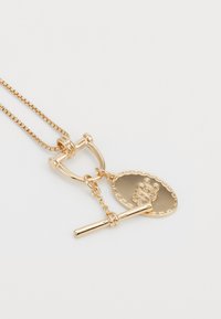 Zlatý náhrdelník s designem řetízků, vybavený přehazovacím zapínáním a oválným přívěskem s vyraženými iniciálami, který zdůrazňuje leštěné povrchy.