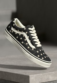 Black canvas sneakers adorned with small white and yellow floral patterns, featuring white laces and a classic rubber sole.