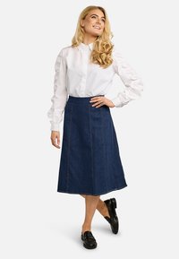 Femme aux longs cheveux blonds porte un chemisier blanc à manches longues, une jupe bleu foncé jusqu'aux genoux et des mocassins noirs, debout avec une main sur la hanche.