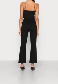 Kobieta ubrana w czarne spodnie z wysokim stanem i szerokimi nogawkami, czarny top na ramiączkach oraz czarne sandały na obcasie, stojąca na jednolitym tle.