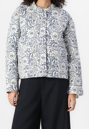 Vrouw die een wit-blauw jasje draagt met een bloemen paisley patroon, met klepzakken aan de voorkant en een zwarte broek.