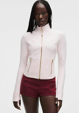 Femme aux longues tresses portant une veste zippée rose clair et un short rouge foncé, debout devant un fond clair uni.