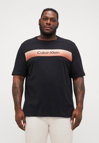 Juodas marškinėliai su trumpomis rankovėmis, turintys rožinės spalvos gradientinę juostą per krūtinę su „Calvin Klein“ tekstu storu šriftu.