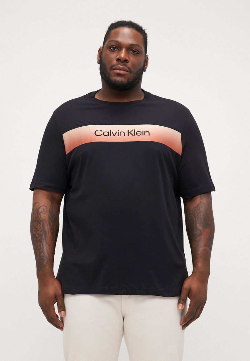 Juodas marškinėliai su trumpomis rankovėmis, turintys rožinės spalvos gradientinę juostą per krūtinę su „Calvin Klein“ tekstu storu šriftu.