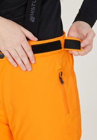 Orange utomhusbyxor med blixtlås-fickor och kardborreband i midjan. Materialet verkar vara hållbart. En del av en svart skjorta är synlig ovanför midjan.