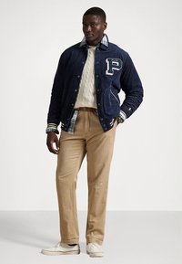 Granatowa bomberka z sztruksu z białymi literami, beżowe spodnie i dziergany sweter. Warstwy obejmują koszulę w kratę oraz casualowe białe buty.