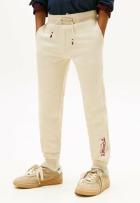 Beige Jogginghosen aus weichem Material, mit einem Kordelzug-Bund, Seitentaschen und einem kleinen roten Logo am linken Bein.