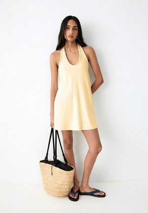 Nainen, jolla on keltainen hihaton mekko ja siniset varvassandaalit, pitelemässä suurta punottua olkilaukkua, jossa on mustat kahvat.