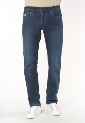Jeans slim fit blu indossati con scarpe da ginnastica bianche su una persona in piedi davanti a uno sfondo uniforme.