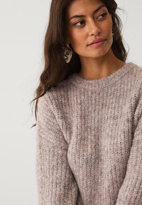 Lys pink strikket sweater med ribbet tekstur og rund halsudskæring. Modelen bærer oversized øreringe, blødt indrammet af langt, løst hår.