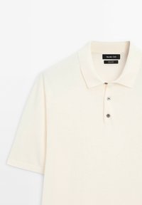 Polo en coton couleur crème avec un col classique, des manches courtes et une patte à trois boutons avec des boutons contrastants foncés.