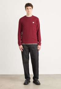 Bordeaux langeftrui met ronde hals en klein logo, gecombineerd met donkere jeans en zwarte schoenen. Soepele textuur, casual pasvorm.