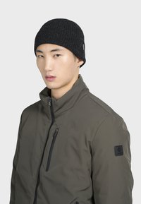 Beanie a coste nera con una vestibilità aderente, abbinato a un giubbotto verde con zip dotato di colletto alto e toppa con logo sulla manica.