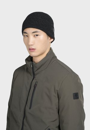 Bonnet côtelé noir à la coupe près du corps, associé à une veste zippée verte avec un col haut et un écusson de marque sur la manche.