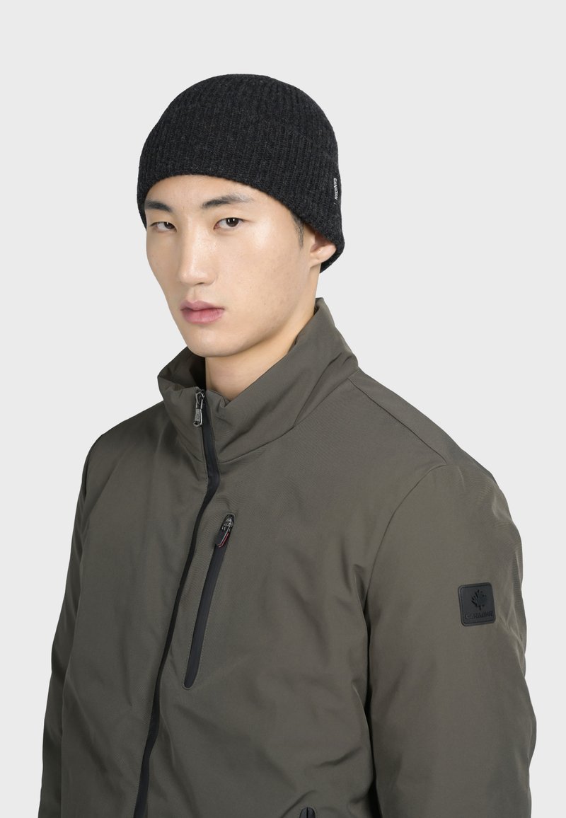 Beanie a coste nera con una vestibilità aderente, abbinato a un giubbotto verde con zip dotato di colletto alto e toppa con logo sulla manica.