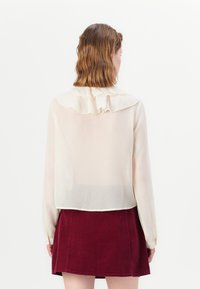 Gjennomsiktig kremfarget bluse med volangkrage, lange ermer og lett cropped design, båret over et burgunder kordfløyelskjørt.