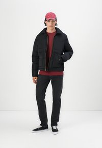 Chaqueta de plumas negra con cuello y dos bolsillos frontales, combinada con una camisa burdeos y jeans oscuros. El modelo lleva una gorra rosa y deportivas negras.