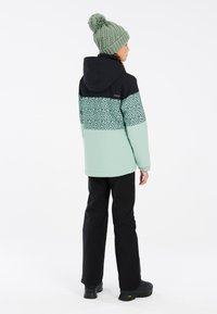 Giacca verde menta con sezione superiore nera e motivo a pois, abbinata a pantaloni neri e un berretto di maglia verde con pompon.