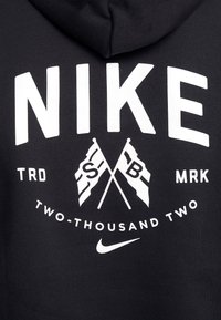 Svart huvtröja med stor vit "NIKE"-text, med två korslagda flaggor, cirkulär grafik och texten "TVÅ-TUSEN TVÅ" nedanför.