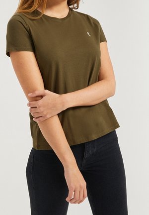 Vrouw draagt een olijfgroen T-shirt met korte mouwen en een klein wit logo op de borst, gecombineerd met een zwarte spijkerbroek, armen gekruist ter hoogte van de taille.