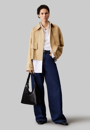 Woman wearing a beige cropped jacket, white shirt, wide-leg dark jeans, black shoes, holding a black handbag, standing against a gray background.