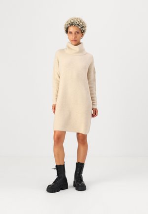Robe pull en tricot beige avec un col roulé haut, des épaules tombantes et une longueur mi-cuisse, associée à des bottines noires à lacets.