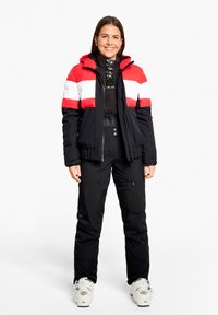 Gepolsterte Skijacke in Schwarz, Rot und Weiß mit Kapuze, kombiniert mit schwarzen wasserdichten Hosen und weißen Skischuhen. Mit Reißverschlüssen und verstellbaren Bündchen.