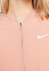 Primer plano de una persona que lleva una camiseta deportiva de color rosa claro con cremallera, con una pequeña textura de puntos y un logo de Nike en blanco en el lado derecho.