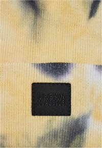Rumena pletena kapa z motivom tie-dye v sivi barvi. Ima črno blago oznako z embossiranim napisom "URBAN CLASSICS." Mehka tekstura.