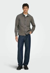 Camisa de botón gris con dos bolsillos en el pecho, combinada con jeans de mezclilla sueltos de color azul oscuro y zapatos negros de cordones. Fondo liso.
