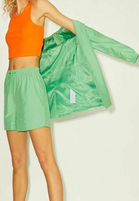 Giacca leggera verde con finitura satinata e pantaloni corti abbinati. Top corto arancione in esposizione, caratterizzato da un design aderente.