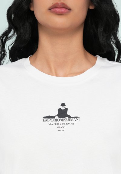 Balts kokvilnas T-krekls ar melnu grafisko logotipu, kurā attēlota sēdoša figūra, ar tekstu "EMPORIO ARMANI VIA BORGONUOVO 11 MILANO SINCE 1981."