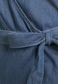 Tkanina dżinsowa w ciemnym niebieskim kolorze, prezentująca fason kopertowy z wiązanym paskiem. Charakteryzuje się miękką fakturą i starannym przeszyciem wzdłuż szwów.