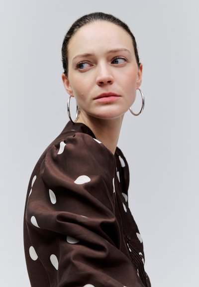 Noor naine siledaks kammitud juustega, suured ringikujulised kõrvakõrvarõngad ja pruun täpiline pluus, vaatab küljele neutraalse ilmega.