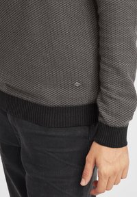 Grå strikket genser med et teksturert mønster, ribbestrikkede mansjetter og hem i en mørkere nyanse, samt en liten logodetalj nær bunnen.