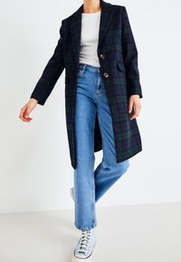 Dlouhý kabát v námořnické modré a zelené kárované vzoru s dvěma knoflíky, výstřihem s výřezy a bočními kapsami, kombinovaný s světle modrými džíny a teniskami.