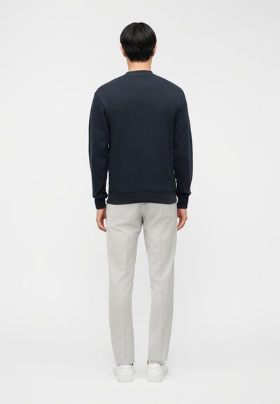 Μπλε ναυτικό βαμβακερό φούτερ με στρογγυλή λαιμόκοψη και ριμπ μανσέτες, συνδυασμένο με ανοιχτό γκρι tailored παντελόνι και λευκά αθλητικά παπούτσια, ορατό από την πίσω πλευρά.