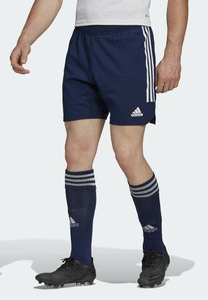 Athlète masculin portant un short de football Adidas bleu marine et des chaussettes assorties avec des crampons noirs, debout dans une position neutre.