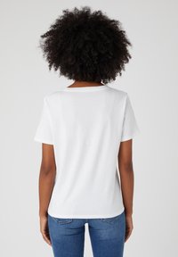 Bílý bavlněný tričko s krátkými rukávy, kulatým výstřihem a volným střihem. Zadní pohled ukazuje hladkou texturu a jednoduchý design, spárované s modrými džínami.